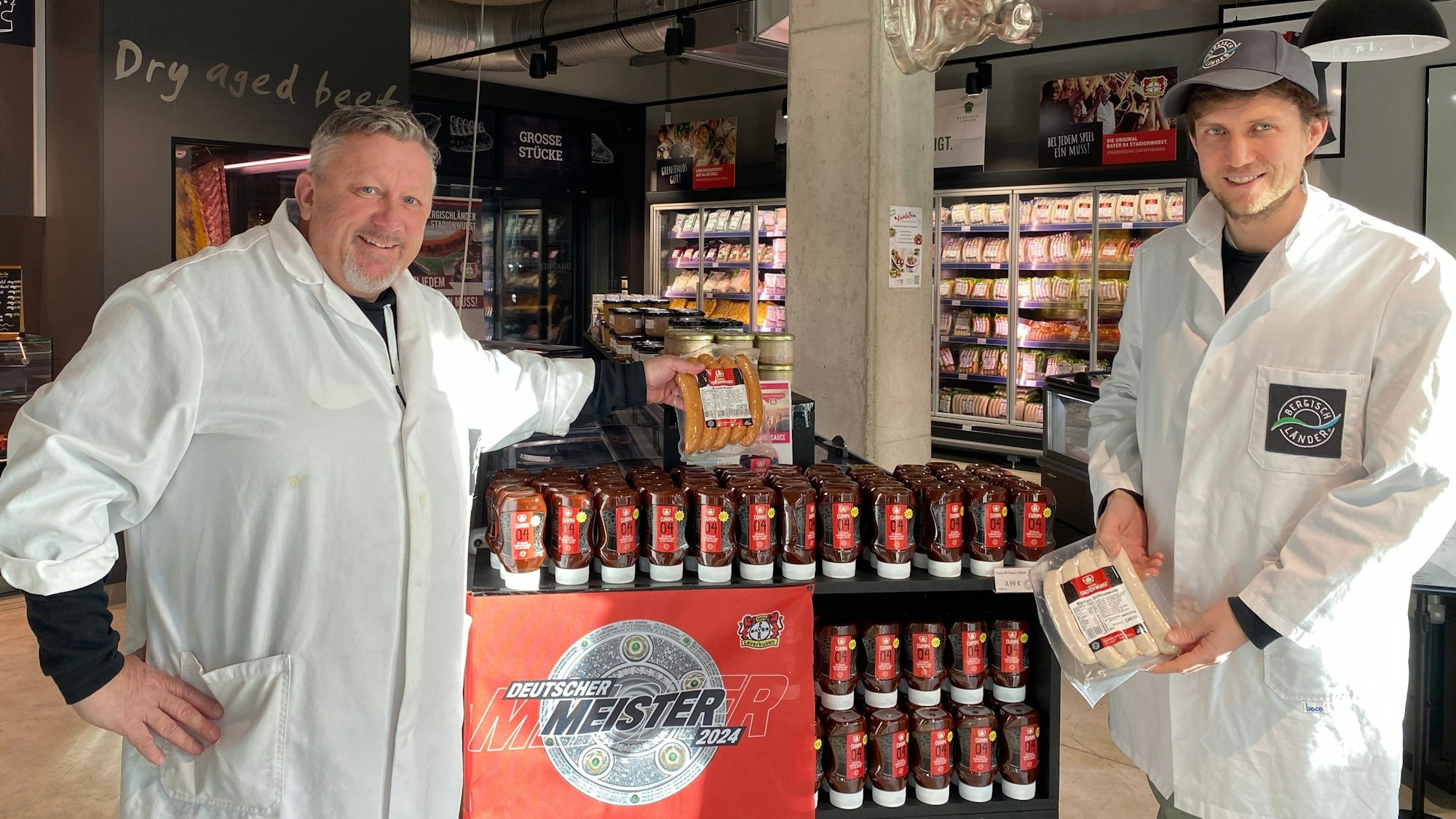 Uwe Nickut und Marcel Schröder stehen in weißen Kittel mit Stadionwurst und Ktakauer in der Hand vor einem Currysoßenstand in dem Markt „Bergischländer“.