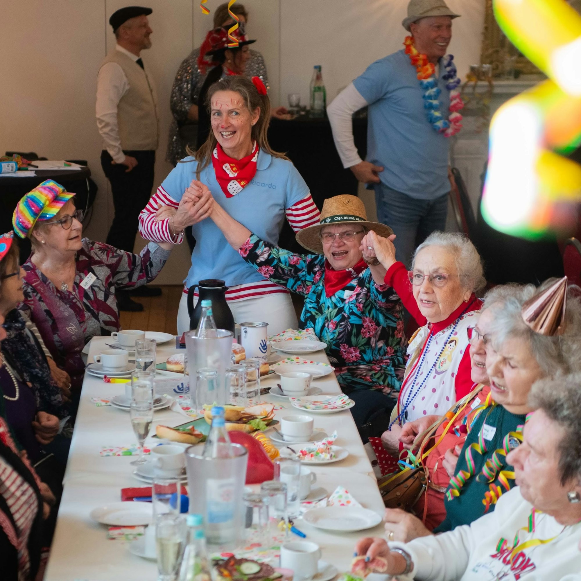 Teilnehmerinnen einer Karnevalsfeier für Senioren sitzen an einem gedeckten Tisch; einige sind kostümiert.