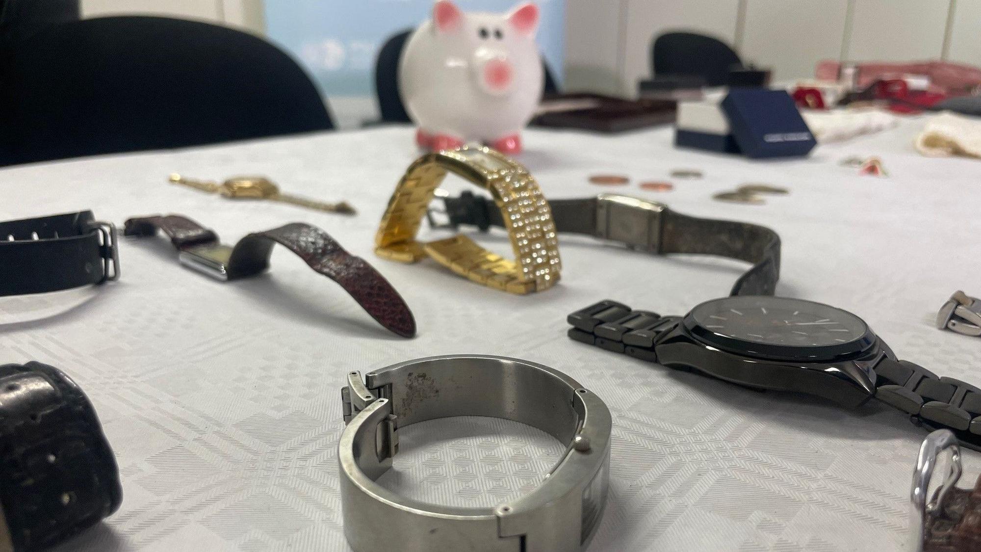 Das Bild zeigt Uhren, die auf einem Tisch liegen. Im Hintergrund steht ein weißes Sparschwein mit rosa Ohren und Füßen.