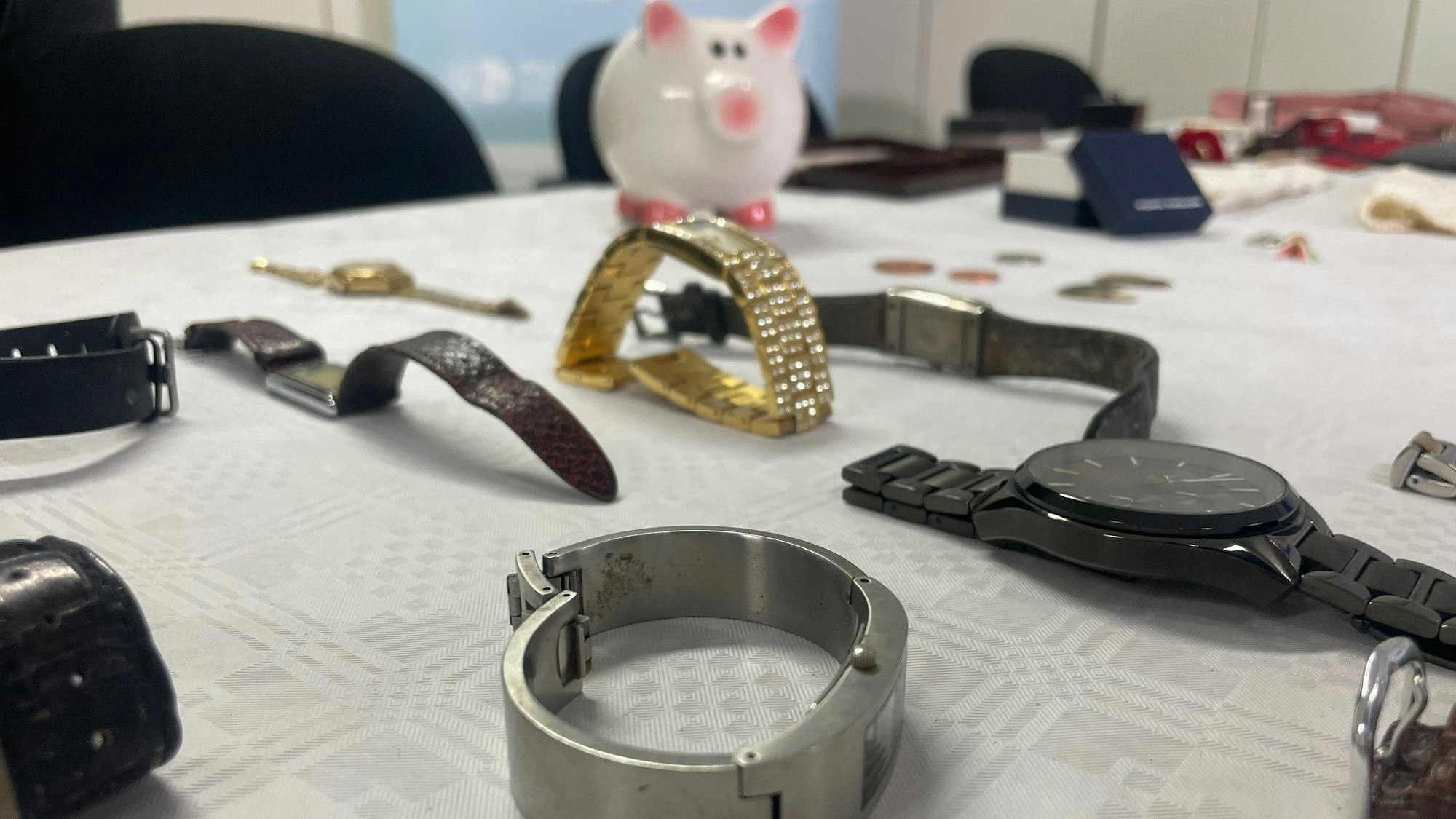 Das Bild zeigt Uhren, die auf einem Tisch liegen. Im Hintergrund steht ein weißes Sparschwein mit rosa Ohren und Füßen.