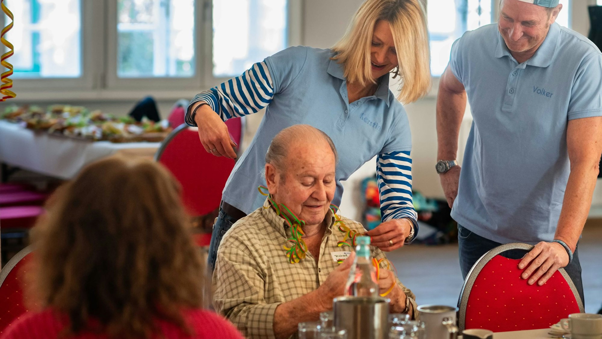 Volker Zimmer (r.) und eine Mitstreiterin aus seinem Verein stehen bei einem älteren Mann, der an einem Tisch sitzt.