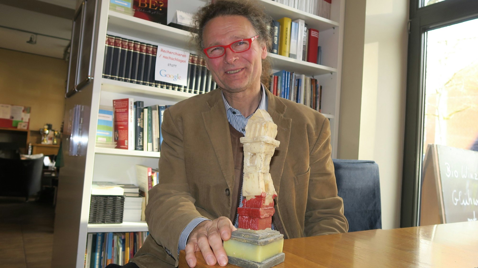Ein Mann mit roter Brille sitzt an einem Tisch vor einem Bücherregal und hält einen „Schrein“ aus Wachs in der Hand.