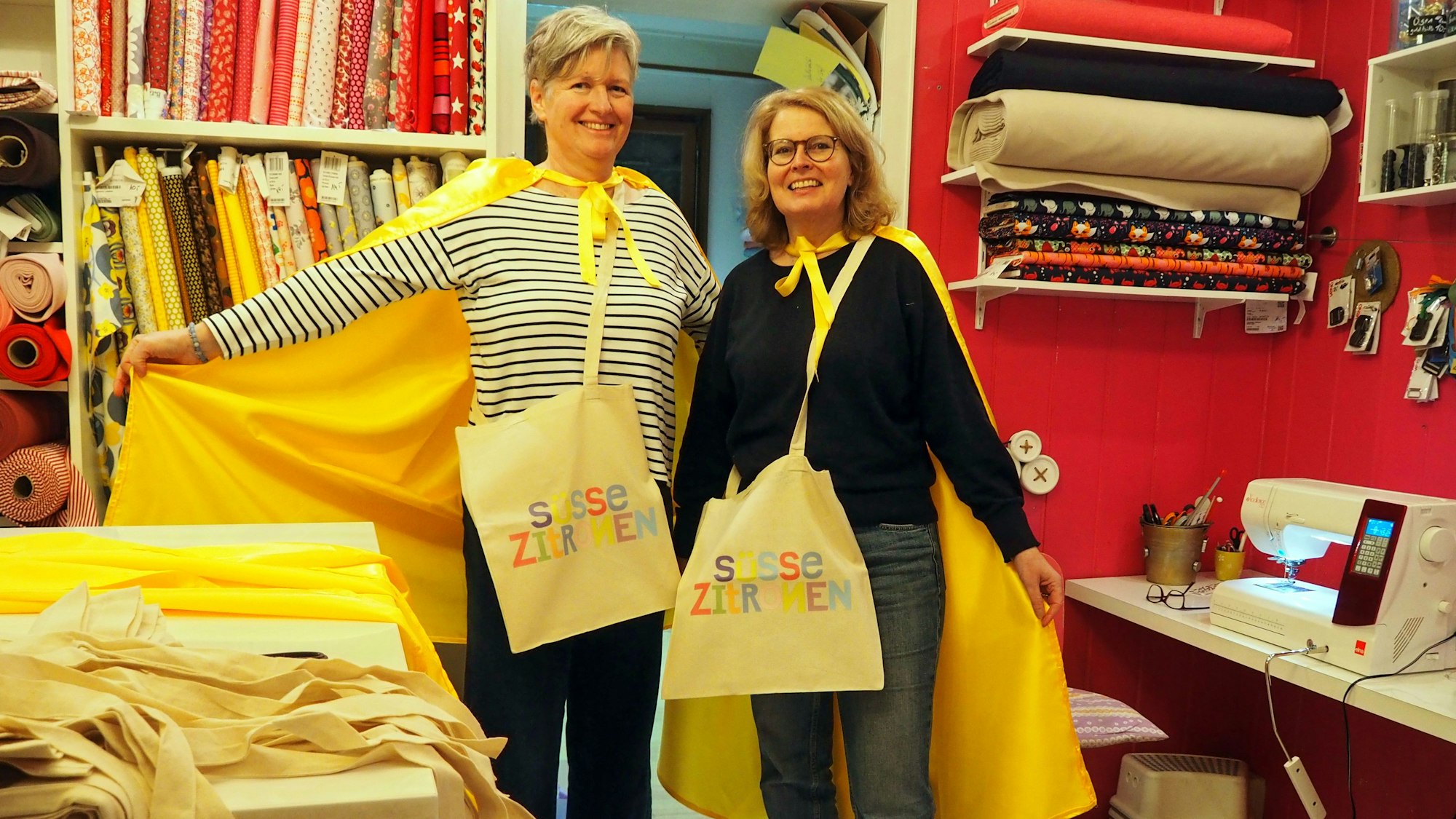 Zwei Frauen mit gelben Umhängen und Jutebeuteln auf denen Süsse Zitronen steht, stehen in einem Nähstudio.