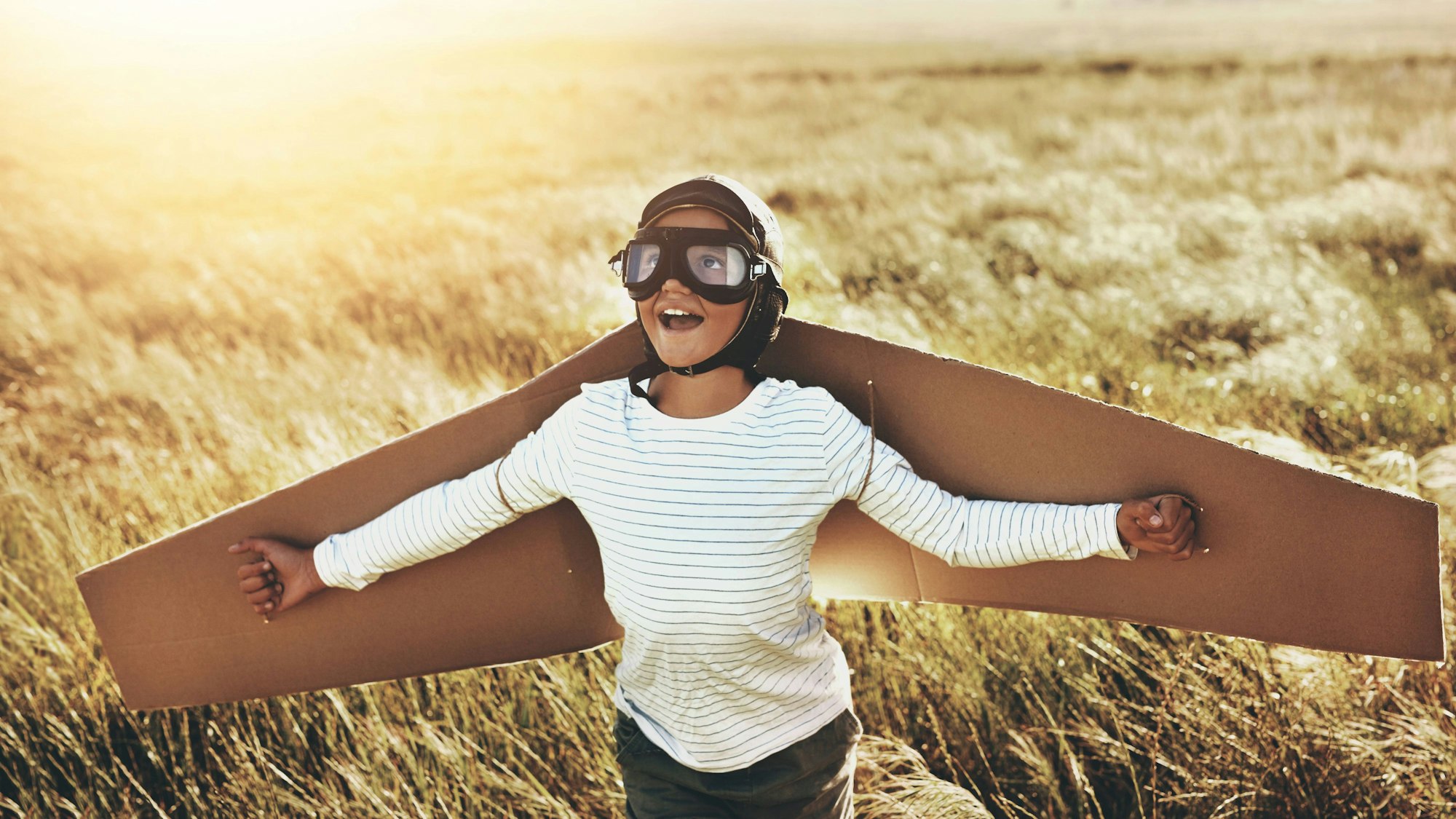 Ein Kind steht mit großen Pappflügeln, einer Pilotenbrille und weit ausgestreckten Armen auf einem Kornfeld