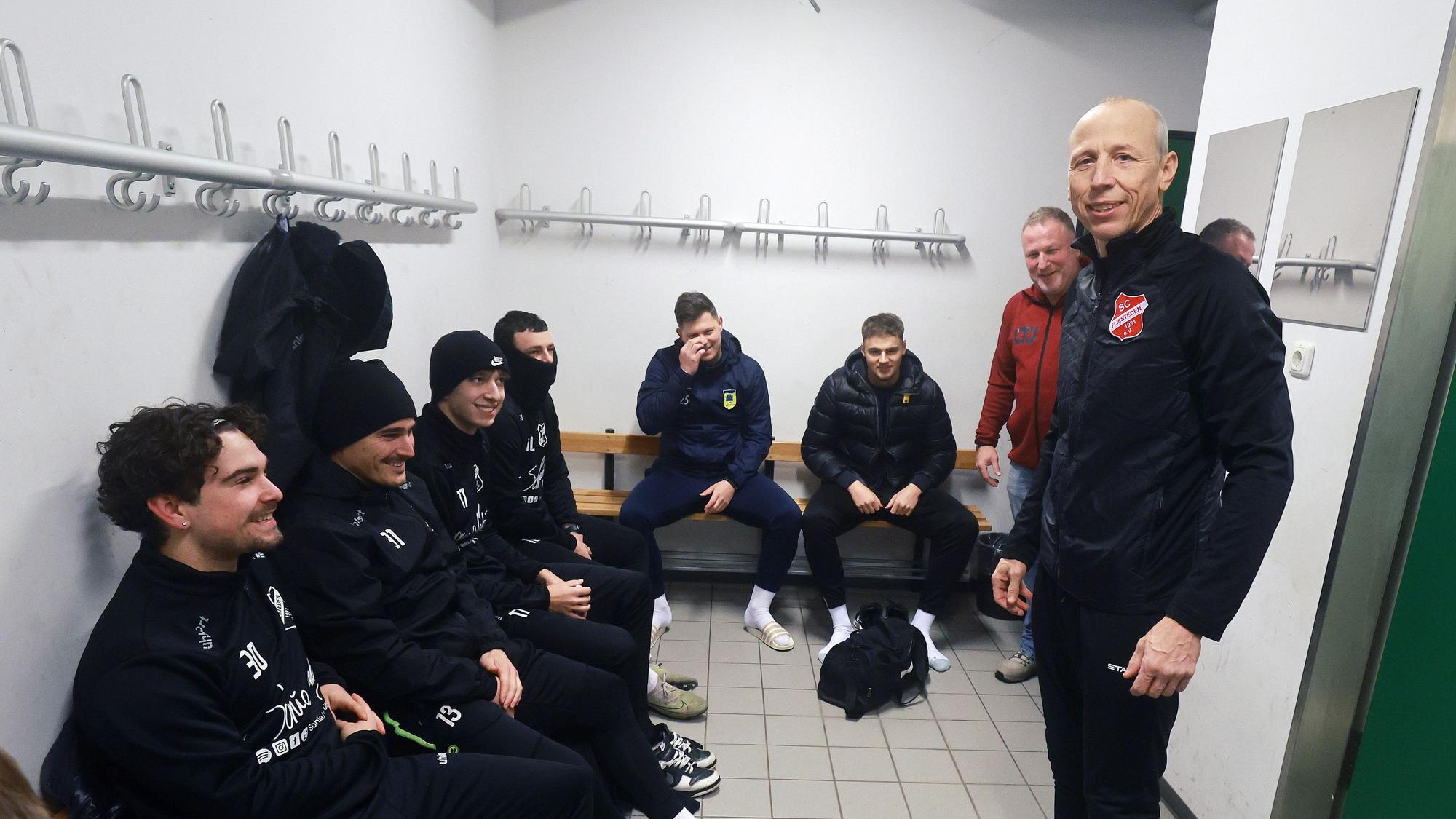 Das Bild zeigt einen Trainer, der in der Kabine vor seinen sitzenden Spielern steht.