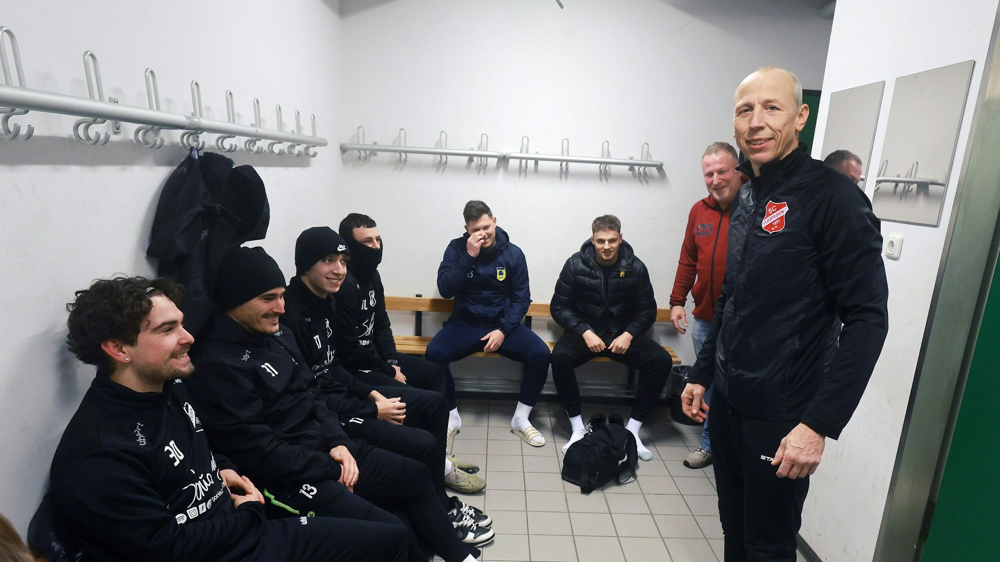 Das Bild zeigt einen Trainer, der in der Kabine vor seinen sitzenden Spielern steht.