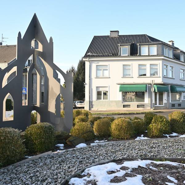 Ein Kreisverkehr mit bepflanzter Mittelinsel, dahinter ein Haus mit Ladenlokal.