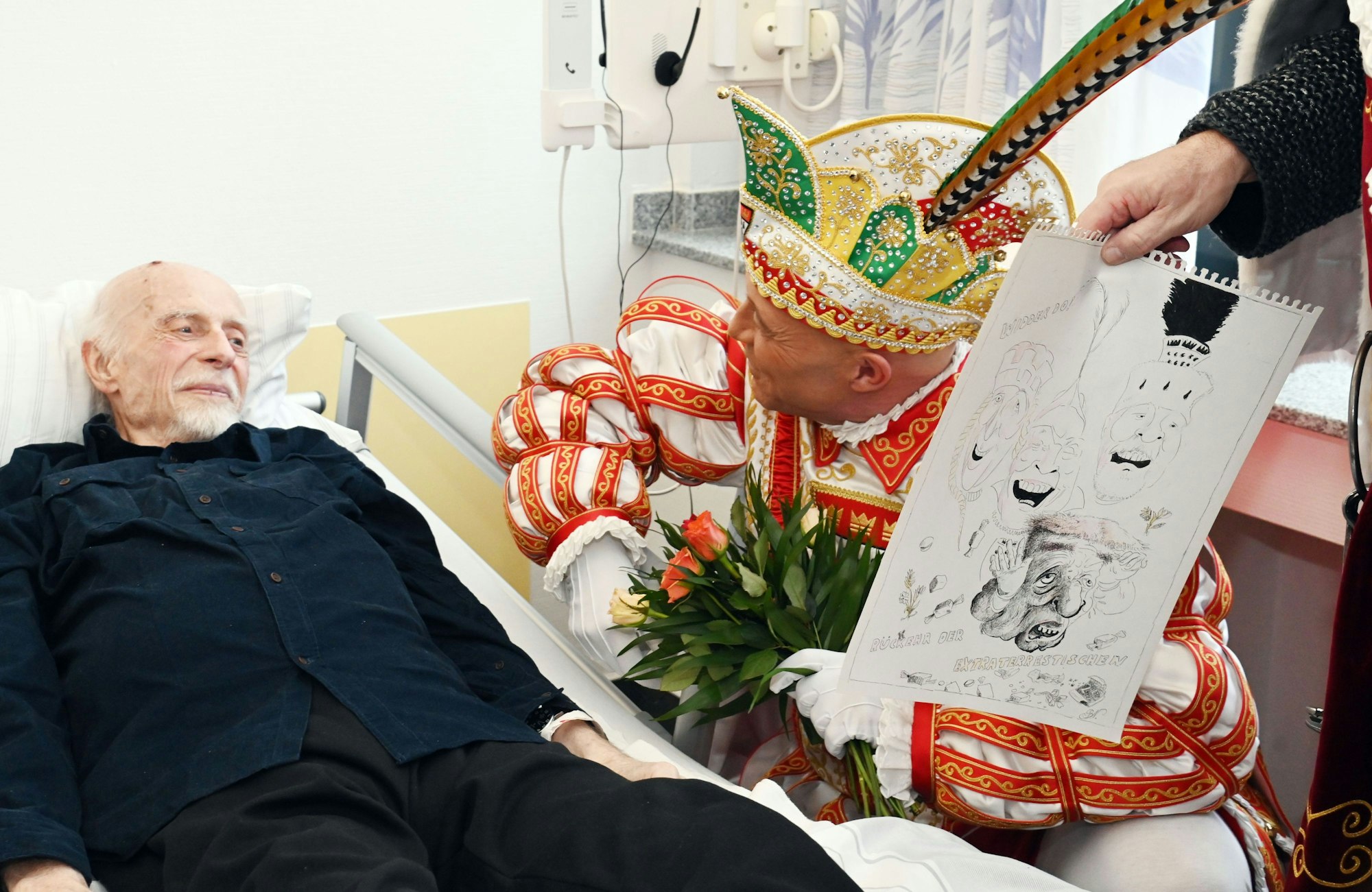 Prinz René I. hockt neben Krankenhausbett, Bauer hält neben ihm eine Comiczeichnung des Dreigestirns.