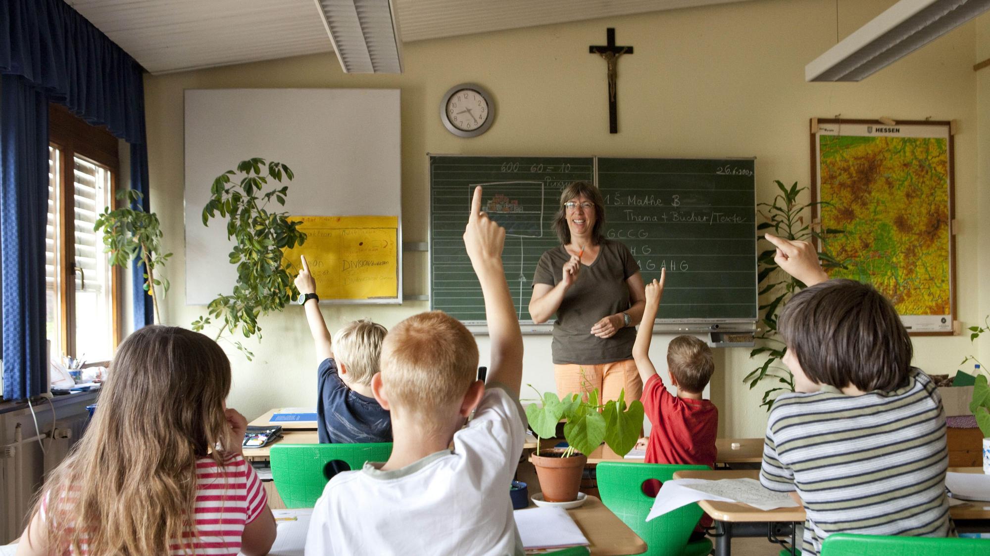 Das Foto zeigt Schüler und eine Lehrerin in einem Klassenzimmer, an der Wand hängt ein Kreuz.
