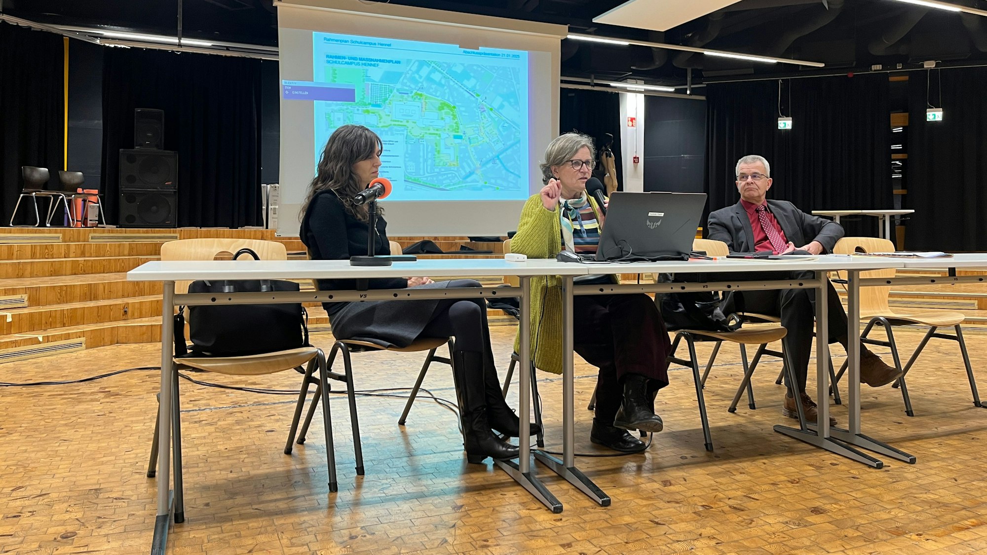 Zwei Frauen und ein Mann sitzen vor einer auf eine Leinwand geworfenen Präsentation.