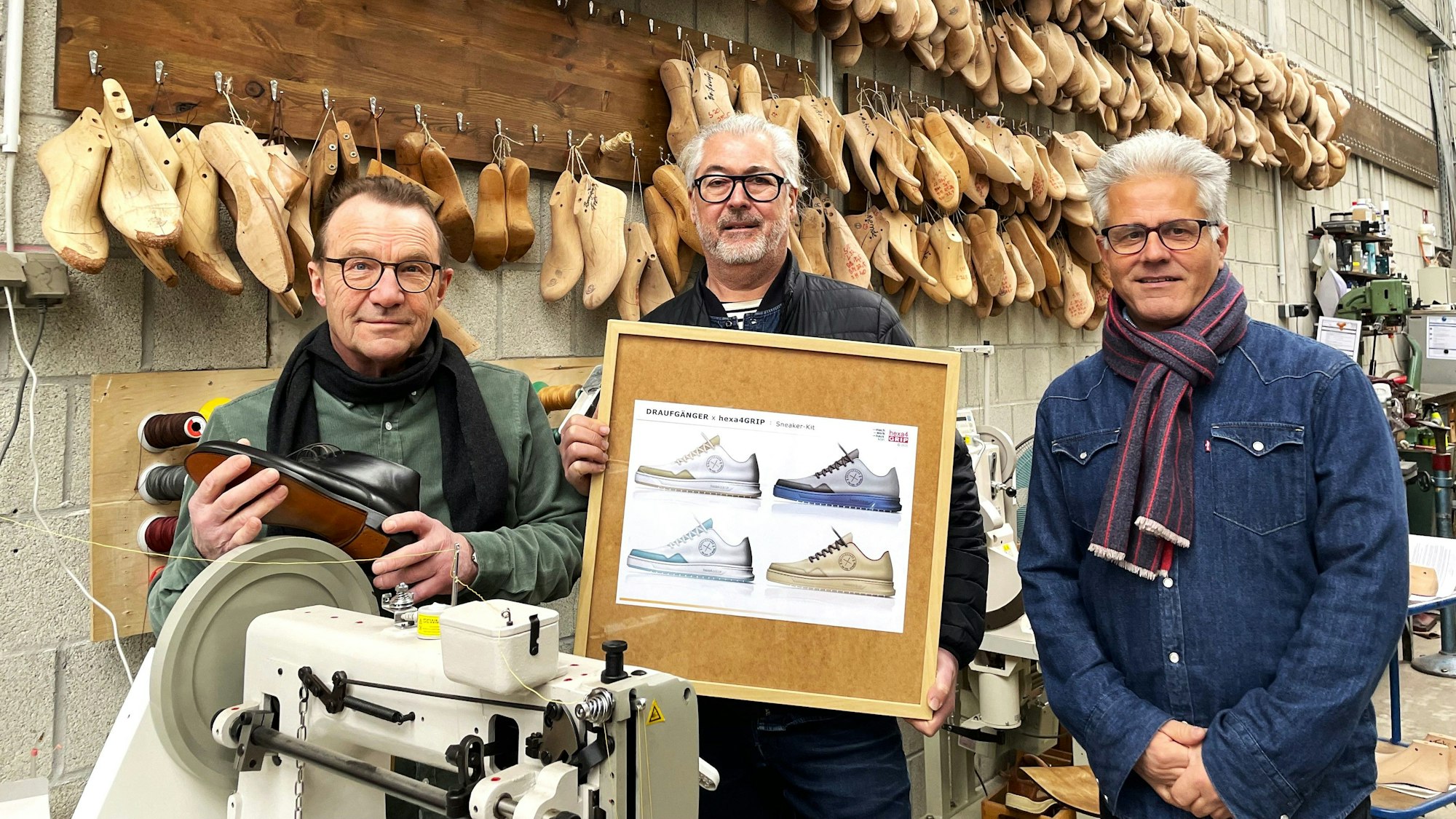 Drei Männer stehen in einer Werkstatt und halten ein Bild in den Händen, auf dem der neue Sneaker zu sehen ist. An der Wand hängen Leisten, Nachbildungen von original Füßen und im Vordergrund eine Nähmaschine.