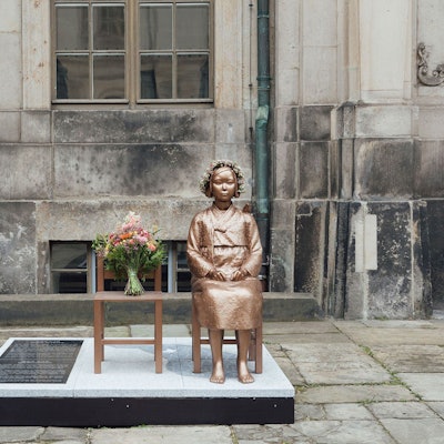 Bronze einer sitzenden Frau neben einem leeren Stuhl.
