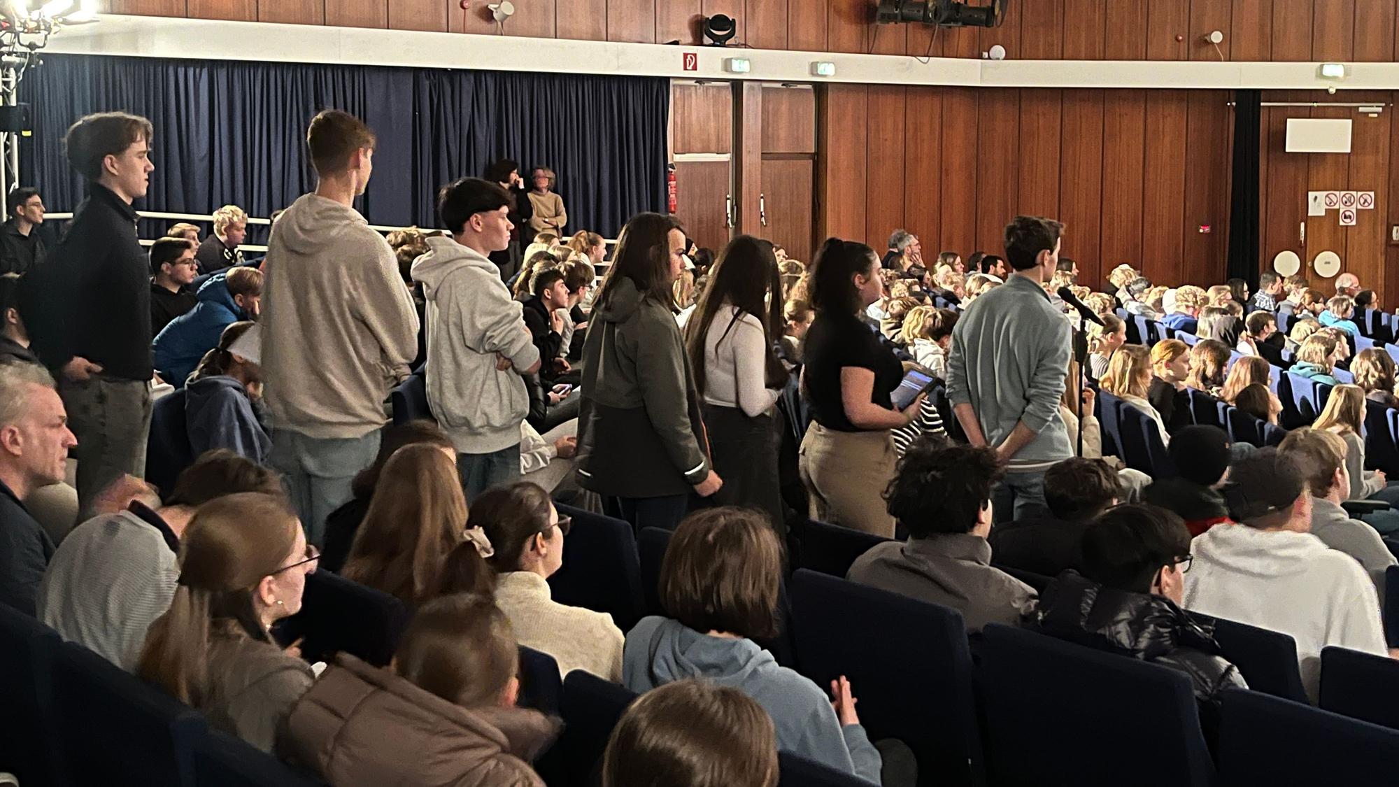 Schüler stehen in einer Aula Schlange vor einem Mikrofon.