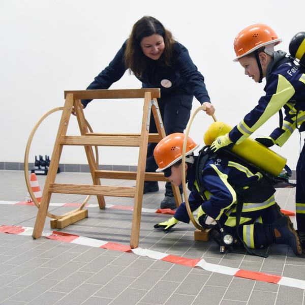 Zwei Kinder in Feuerwehruniform durchlaufen unter Anleitung einen Hindernisparcours.