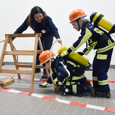 Zwei Kinder in Feuerwehruniform durchlaufen unter Anleitung einen Hindernisparcours.