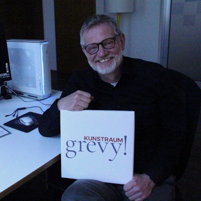 Ein Mann sitzt an einem Schreibtisch, er hält ein Schild, auf dem Kunstraum Grevy steht, auf dem Computer-Monitor ist zur Hälfte ein Zebra zu sehen.
