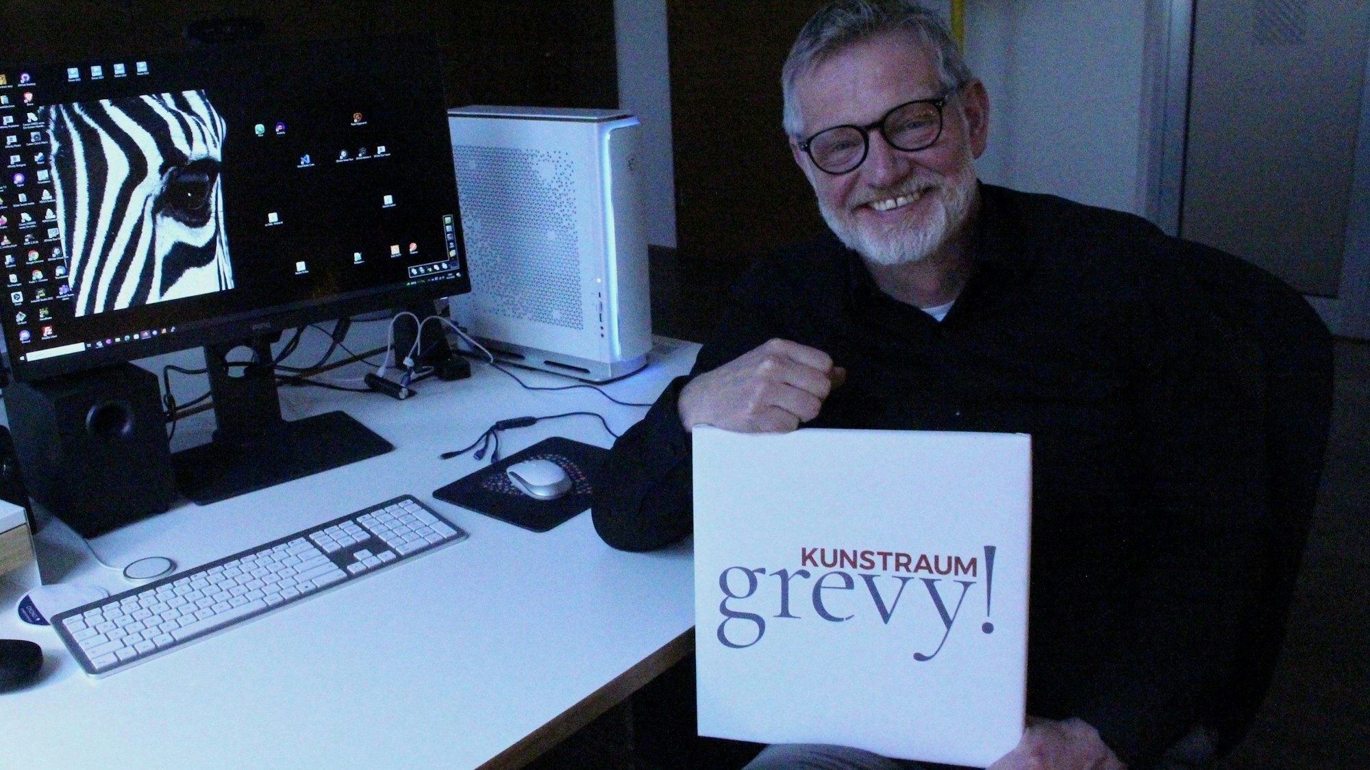 Ein Mann sitzt an einem Schreibtisch, er hält ein Schild, auf dem Kunstraum Grevy steht, auf dem Computer-Monitor ist zur Hälfte ein Zebra zu sehen.