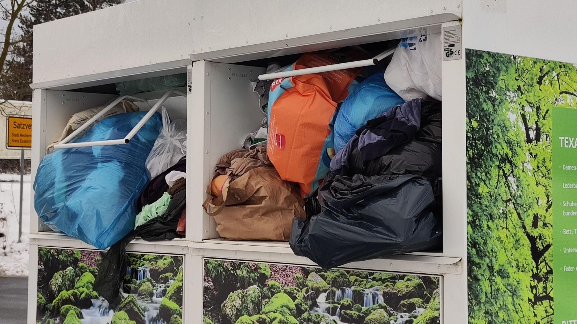 Aus einem Altkleidercontainer quellen Plastiksäcke und Tüten.