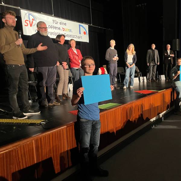 Sechs Politiker und zwei Schüler stehen auf einer Bühne, drei Schüler halten Plakate mit Fragen hoch.