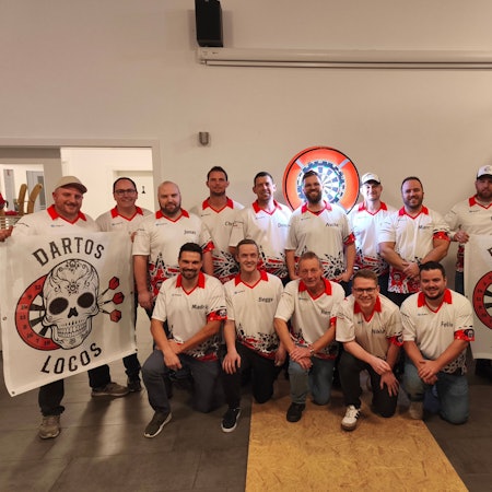 Die Spieler der Dartos Locos aus Billig posieren für die Kamera, links und rechts halten sie Fahnen mit dem Teamlogo hoch. Im Hintergrund ist eine beleuchtete Darts-Scheibe zu erkennen.