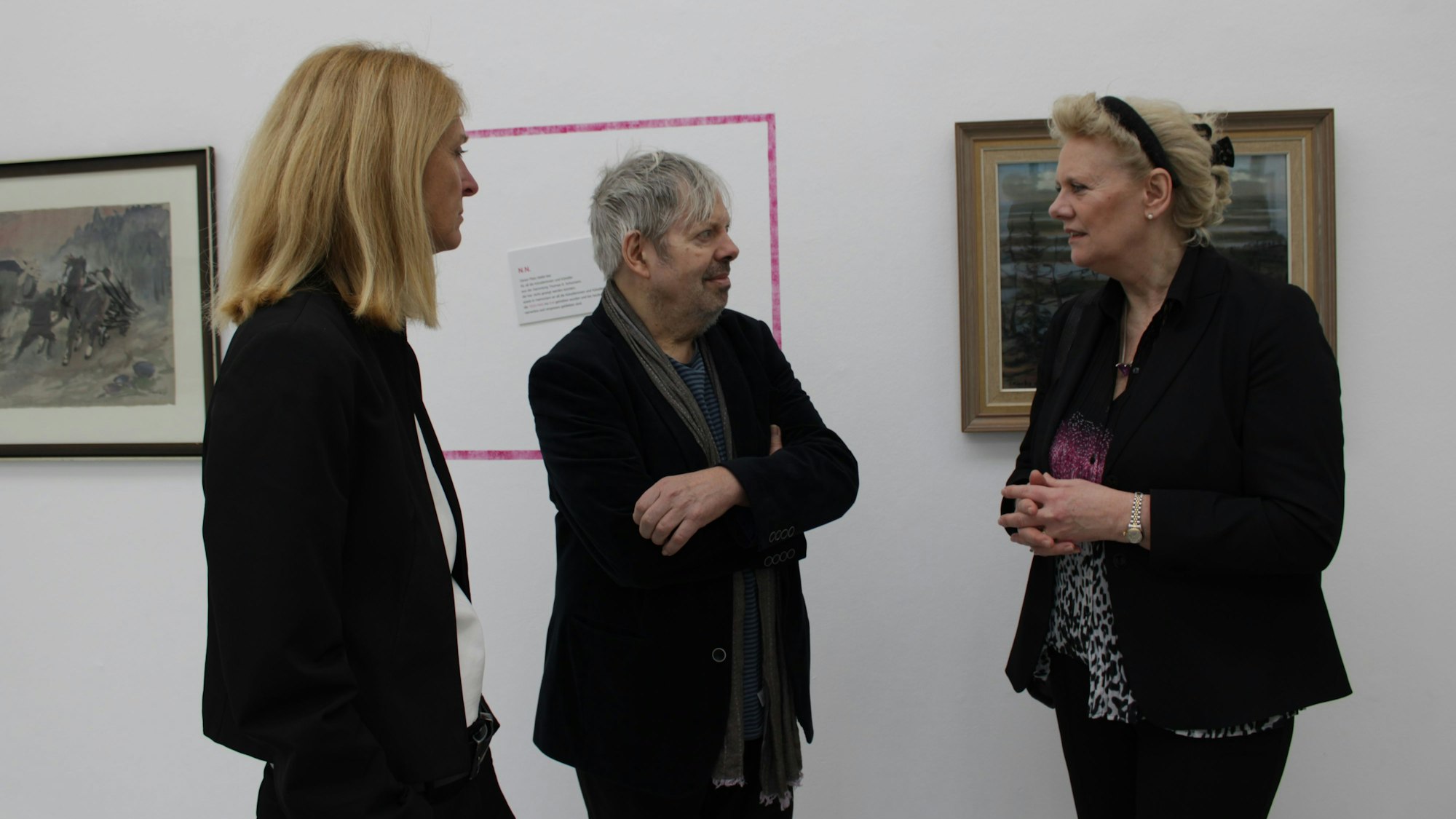 Zwei Frauen und ein Mann stehen vor zwei Bilderrahmen, dazwischen ist ein leerer Rahmen an die Wand gemalt.