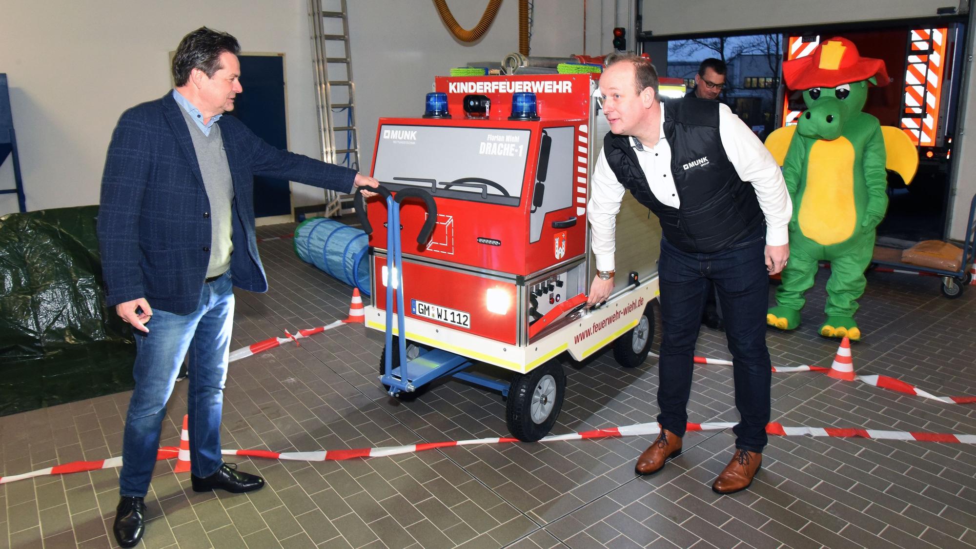Zwei Männer schieben ein gebautes Feuerwehrauto in Miniaturformat auf einem Rollwagen in ein Feuerwehr-Gerätehaus.