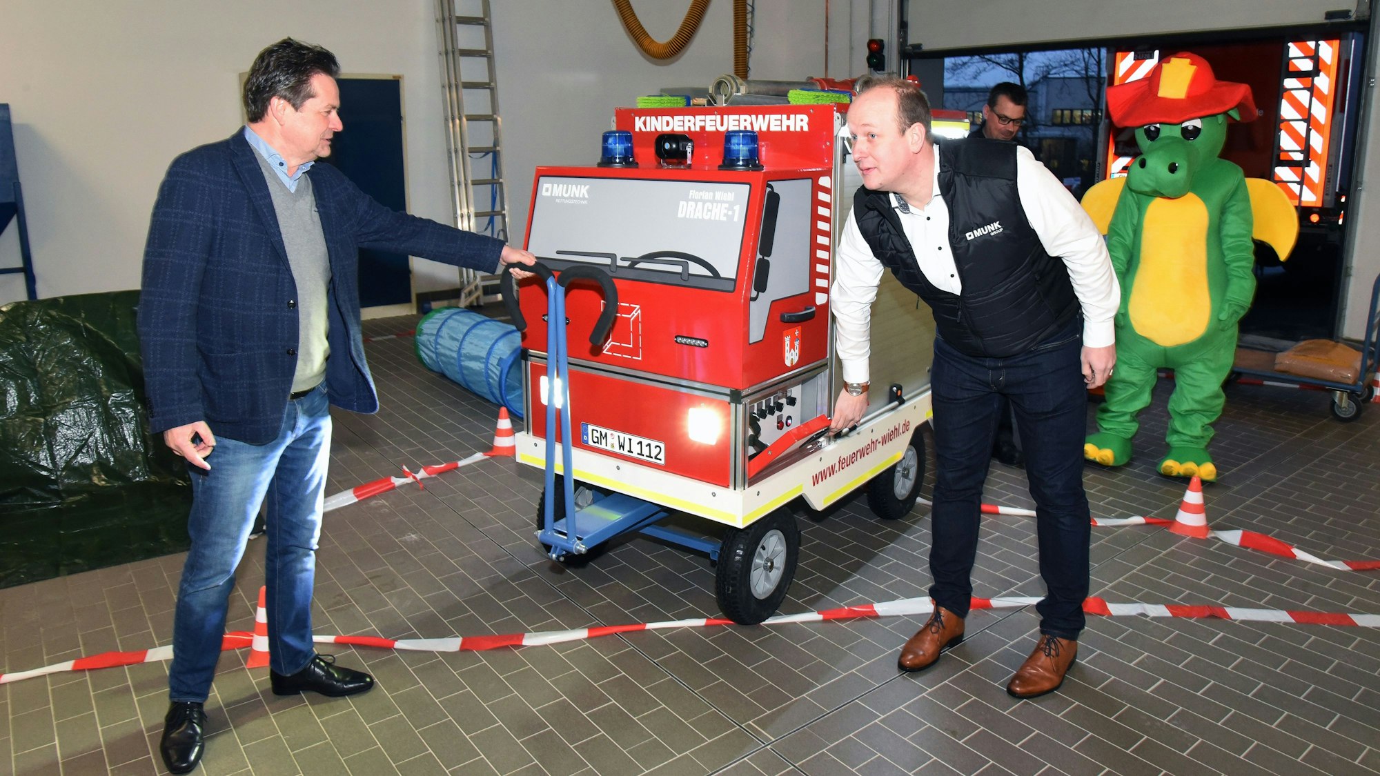 Zwei Männer schieben ein gebautes Feuerwehrauto in Miniaturformat auf einem Rollwagen in ein Feuerwehr-Gerätehaus.