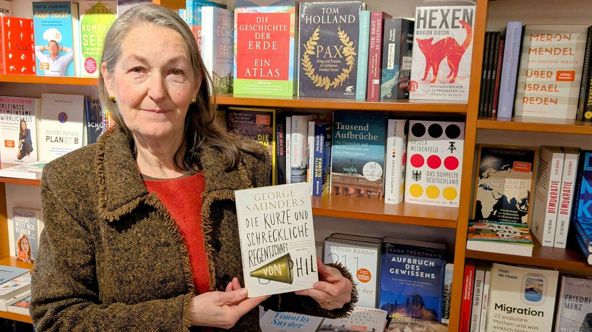 Iris Pütz steht vor einem Bücherregal und präsentiert das Buch „Die kurze und schreckliche Regentschaft von Phil“ von George Saunders.