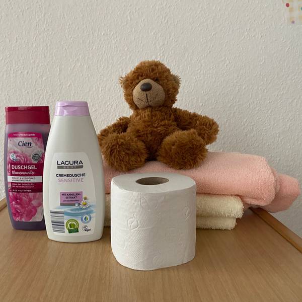Duschgel, eine Rolle Toilettenpapier, zwei Handtücher und ein Teddybär sind auf einem Tisch aufgestellt.