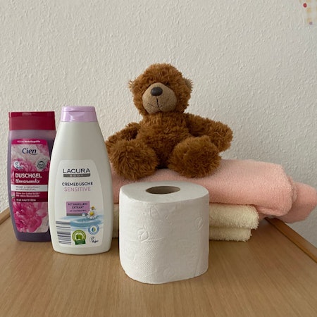 Duschgel, eine Rolle Toilettenpapier, zwei Handtücher und ein Teddybär sind auf einem Tisch aufgestellt.
