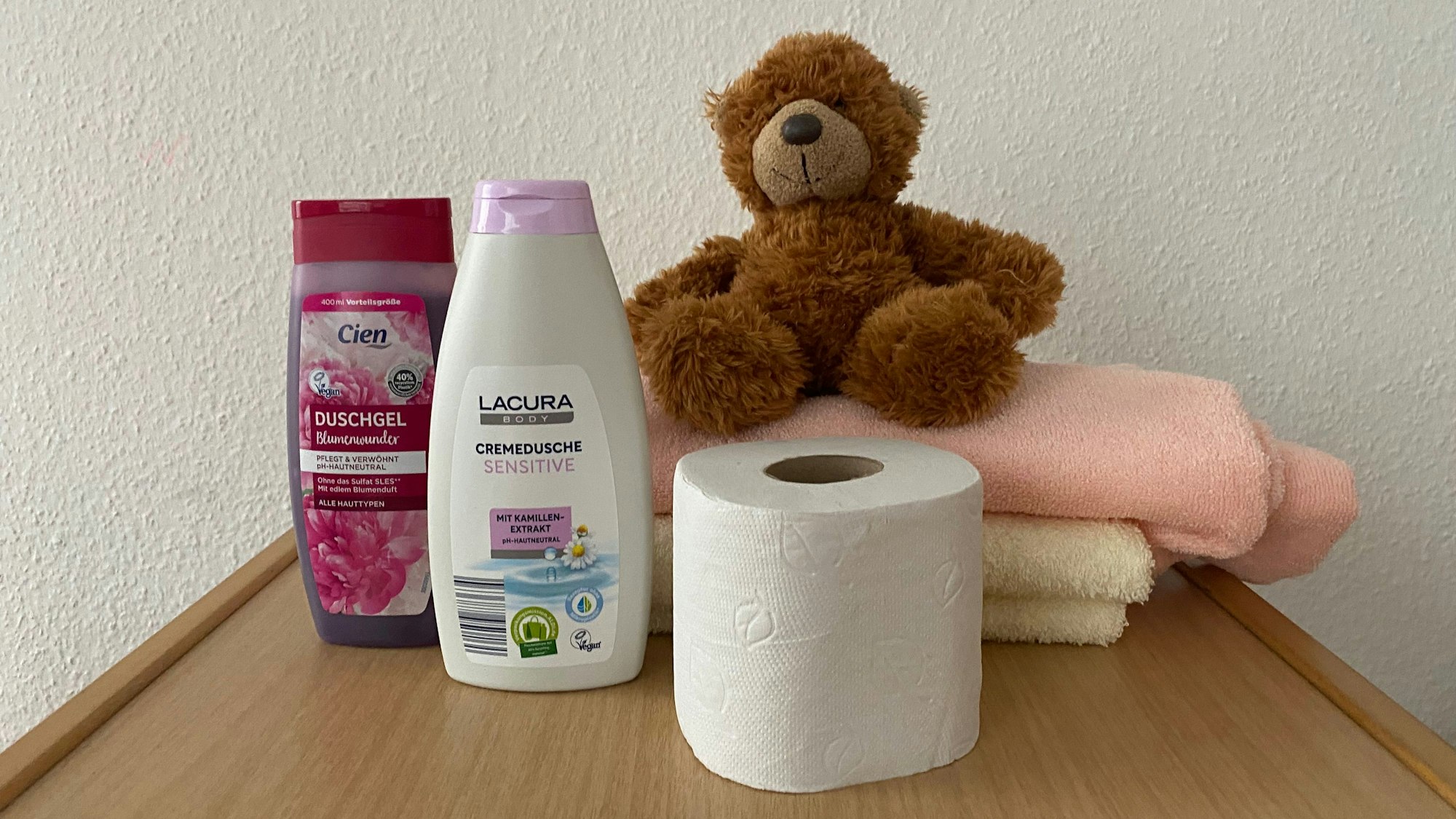 Duschgel, eine Rolle Toilettenpapier, zwei Handtücher und ein Teddybär sind auf einem Tisch aufgestellt.