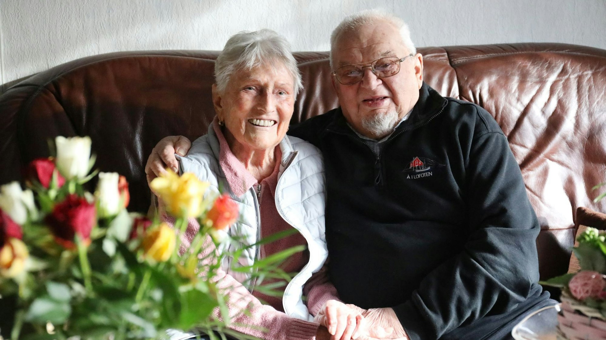 Ein Ehepaar sitzt nebeneinander auf einem Sofa, am Rand steht ein bunter Blumenstrauß.