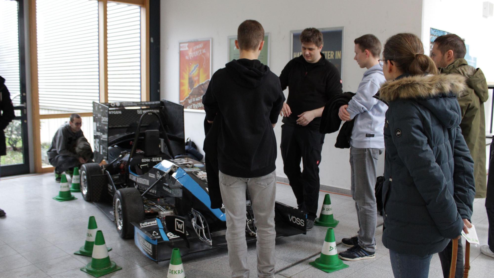 Mehrere Schülerinnen und Schüler stehen vor einem Rennwagen im Flur einer Hochschule, ein Student steht daneben und erzählt etwas.