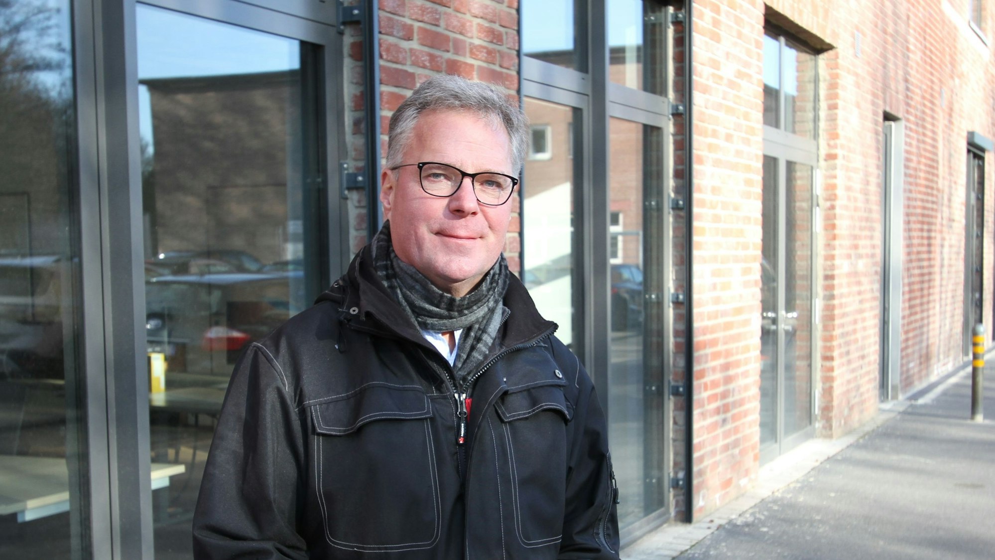 Ein Mann in einer schwarzen Jacke vor einer Glas- und Backsteinfassade.