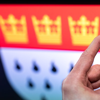 Jemand hält die Testkassette eines Selbst-Schnelltests auf das Coronavirus in die Kamera, im Hintergrund ist das Wappen der Stadt Köln zu sehen.