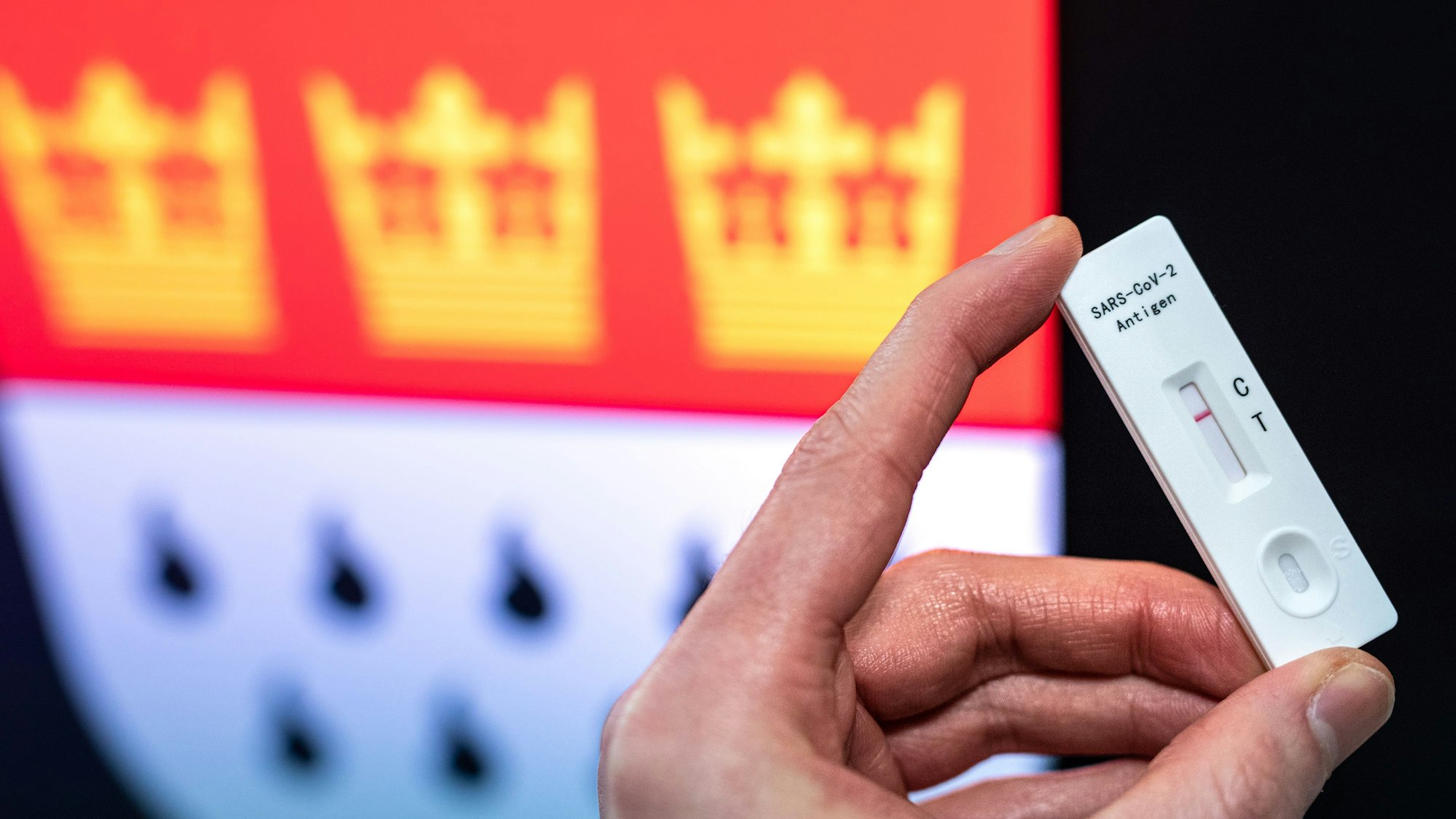 Jemand hält die Testkassette eines Selbst-Schnelltests auf das Coronavirus in die Kamera, im Hintergrund ist das Wappen der Stadt Köln zu sehen.