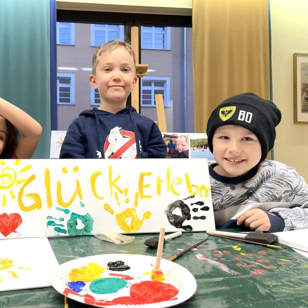 Drei Kinder sitzen an einem Mal-Tisch und halten das selbst gemalte Plakat mit der Aufschrift Glück erleben und den bunten Farb-Abdrücken ihrer Hände in die Kamera.