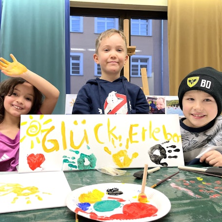 Drei Kinder sitzen an einem Mal-Tisch und halten das selbst gemalte Plakat mit der Aufschrift Glück erleben und den bunten Farb-Abdrücken ihrer Hände in die Kamera.