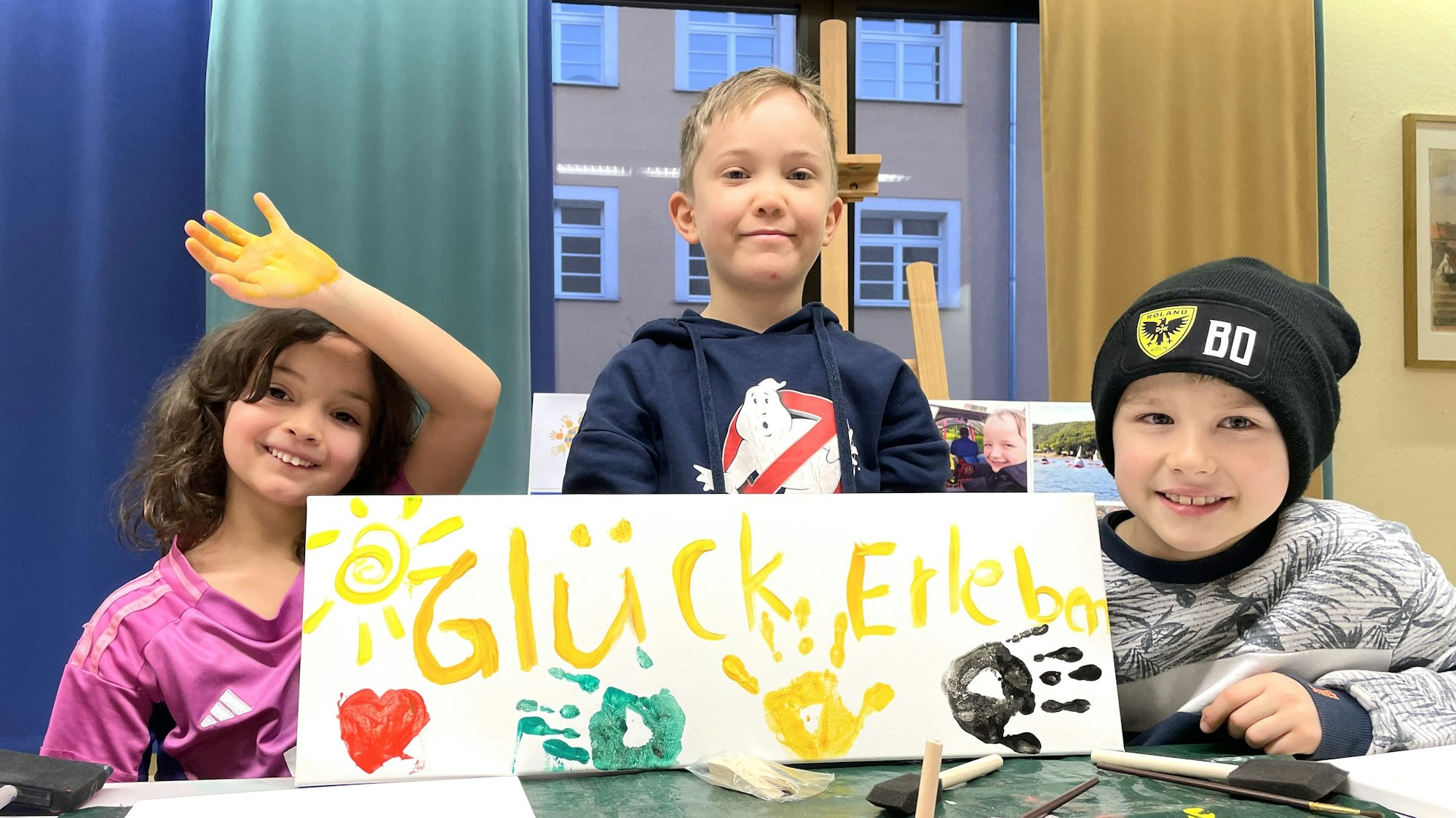 Drei Kinder sitzen an einem Mal-Tisch und halten das selbst gemalte Plakat mit der Aufschrift Glück erleben und den bunten Farb-Abdrücken ihrer Hände in die Kamera.