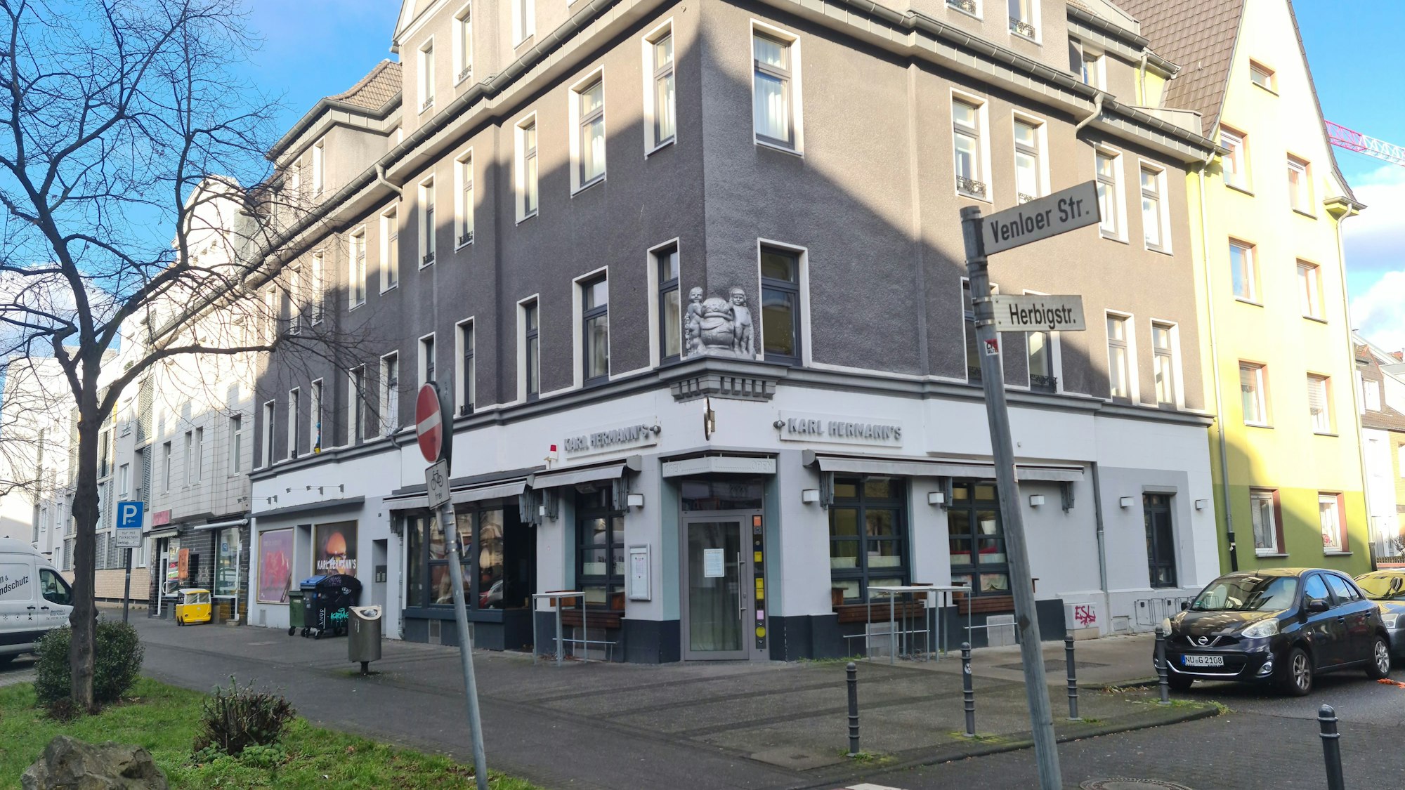 Blick auf ein stattliches Eckhaus mit grauer Fassade, in dessen Erdgeschoss ein Restaurant ist.