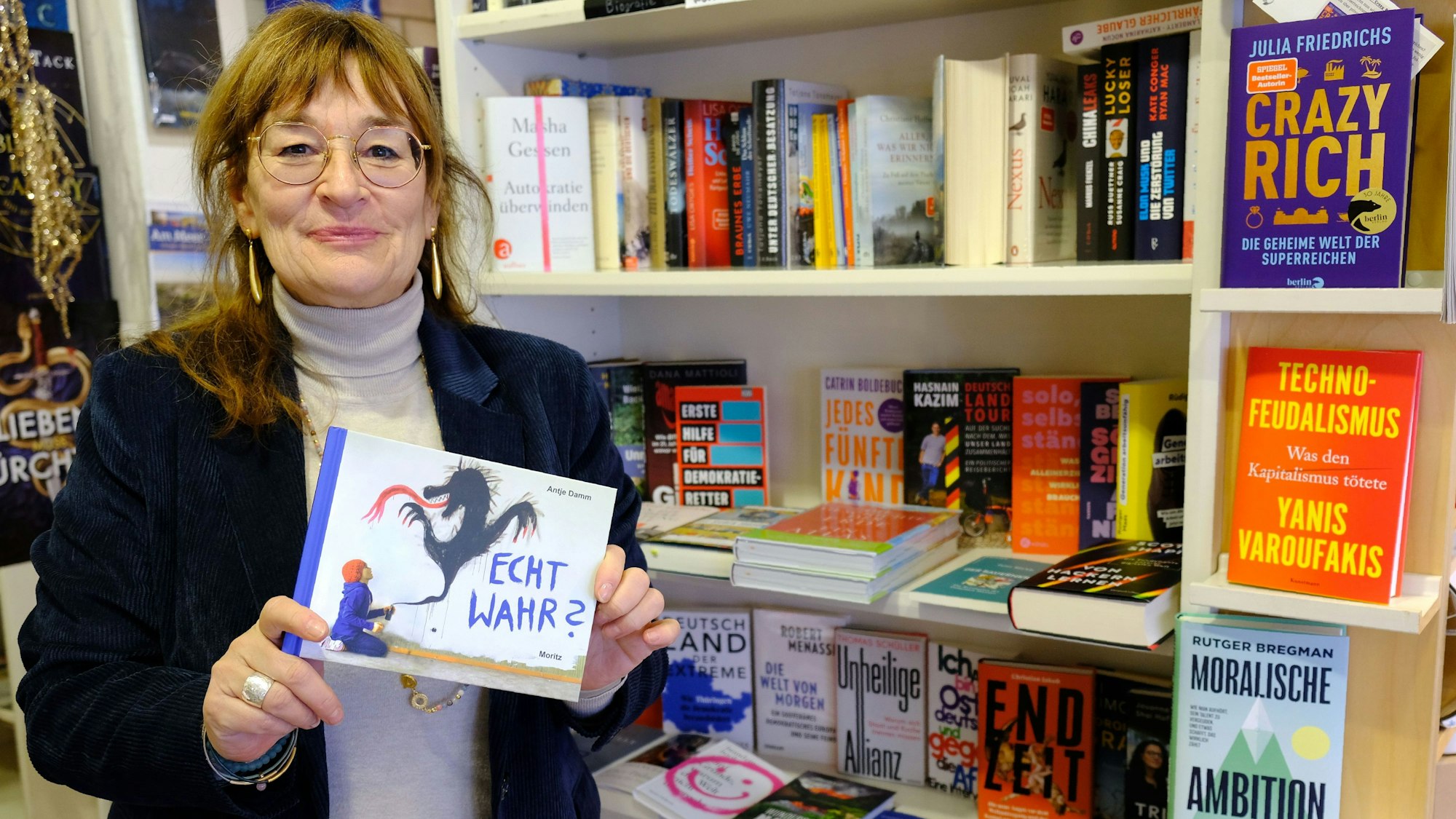 Renate Elsen steht mit einem Exemplar des Buchs „Echt wahr? – 52 Gelegenheiten, sich über Lüge und Wahrheit zu unterhalten“ von Antje Damm vor einem Bücherregal in ihrer Buchhandlung.
