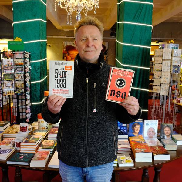 Josef Mütter steht mit zwei Büchern in den Händen in seiner Buchhandlung.