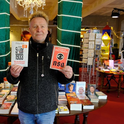 Josef Mütter steht mit zwei Büchern in den Händen in seiner Buchhandlung.