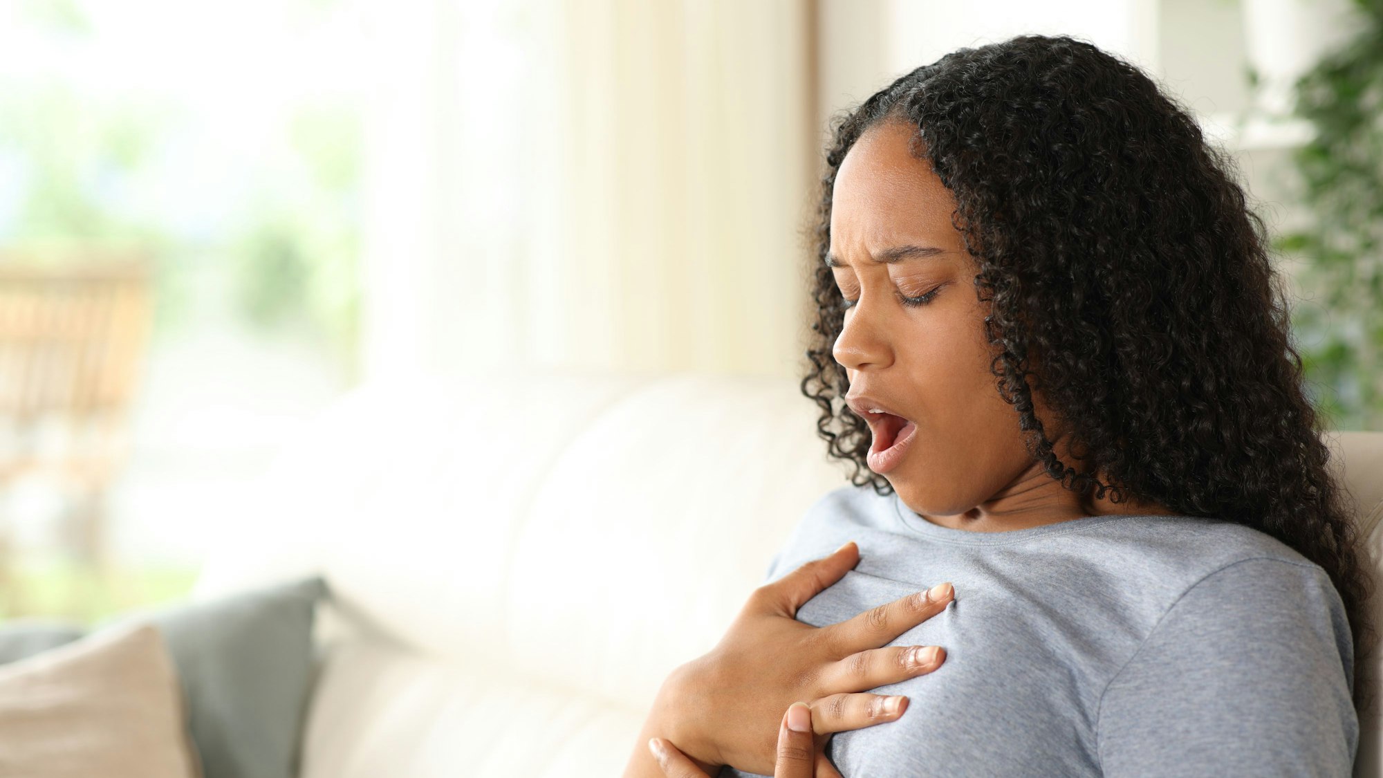 Eine Frau sitzt auf dem Sofa, hält ihre Hände auf ihre Brust und versucht, Luft zu holen.