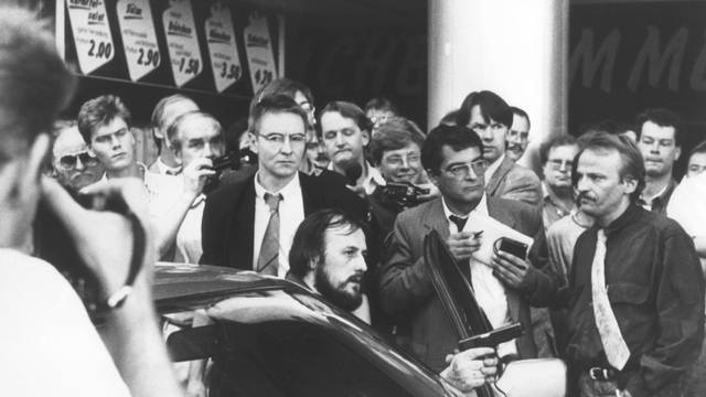 Mit einer Waffe in der Hand fordert der am Steuer des Fluchtfahrzeugs sitzende Hans-Jürgen Rösner in der Kölner Innenstadt die umstehenden Journalisten und Passanten auf, den Weg frei zu machen. Links hinter Rösner steht der Journalist Udo Röbel.