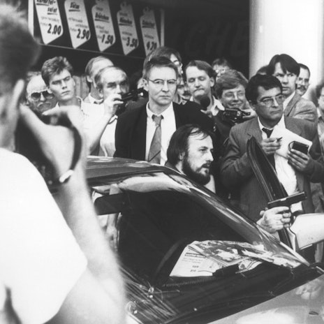 Mit einer Waffe in der Hand fordert der am Steuer des Fluchtfahrzeugs sitzende Hans-Jürgen Rösner in der Kölner Innenstadt die umstehenden Journalisten und Passanten auf, den Weg frei zu machen. Links hinter Rösner steht der Journalist Udo Röbel.