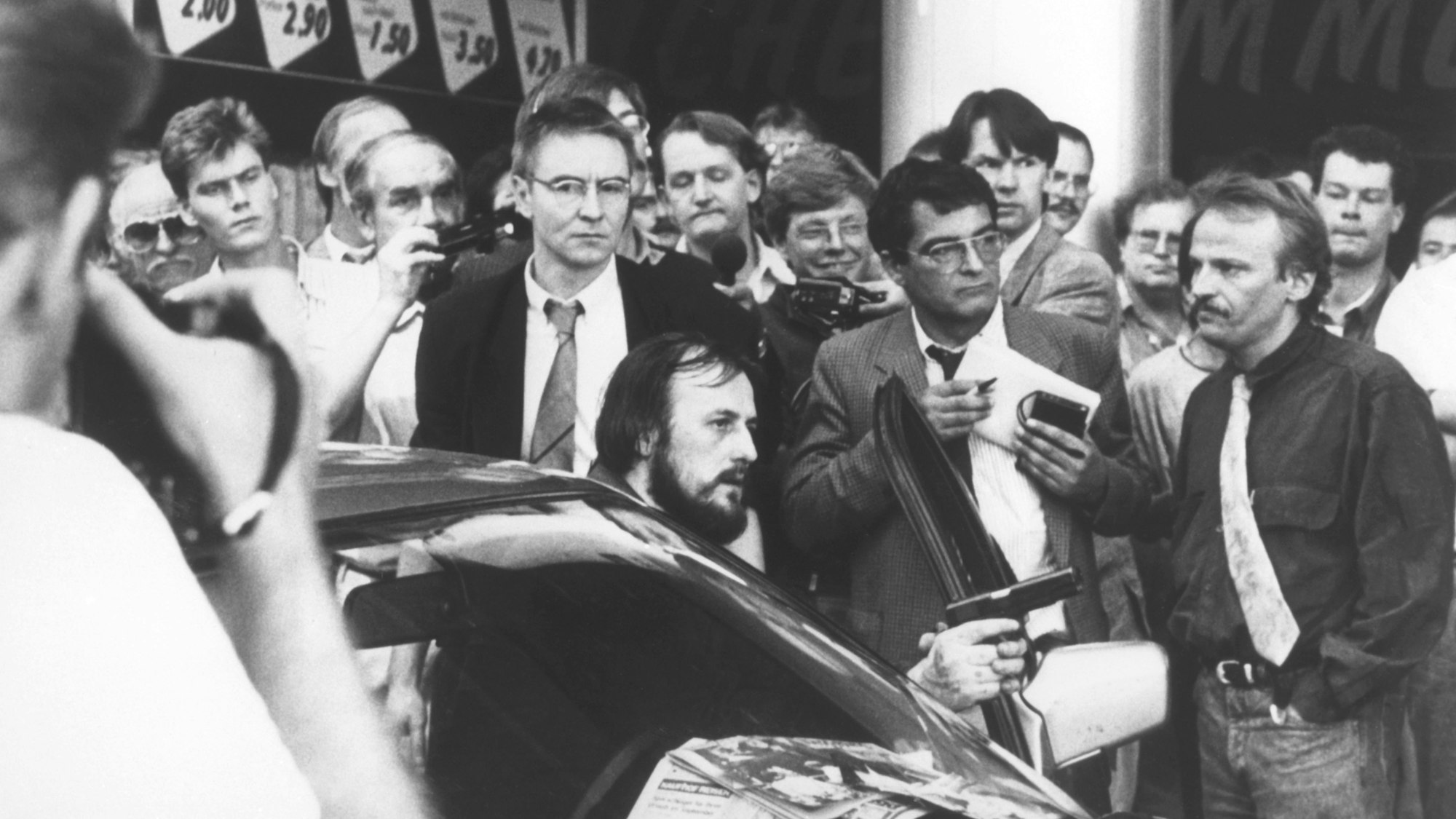 Mit einer Waffe in der Hand fordert der am Steuer des Fluchtfahrzeugs sitzende Hans-Jürgen Rösner in der Kölner Innenstadt die umstehenden Journalisten und Passanten auf, den Weg frei zu machen. Links hinter Rösner steht der Journalist Udo Röbel.