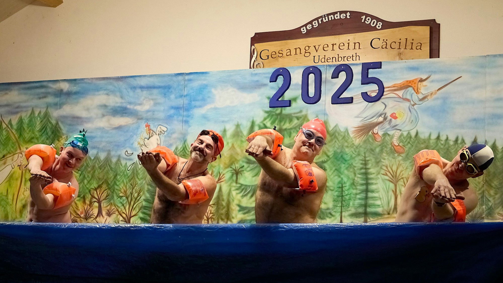 Vier Männer mit bloßem Oberkörper und Schwimmflügeln zeigen vor einem Bühnenbild Bewegungen wie beim Synchronschwimmen.