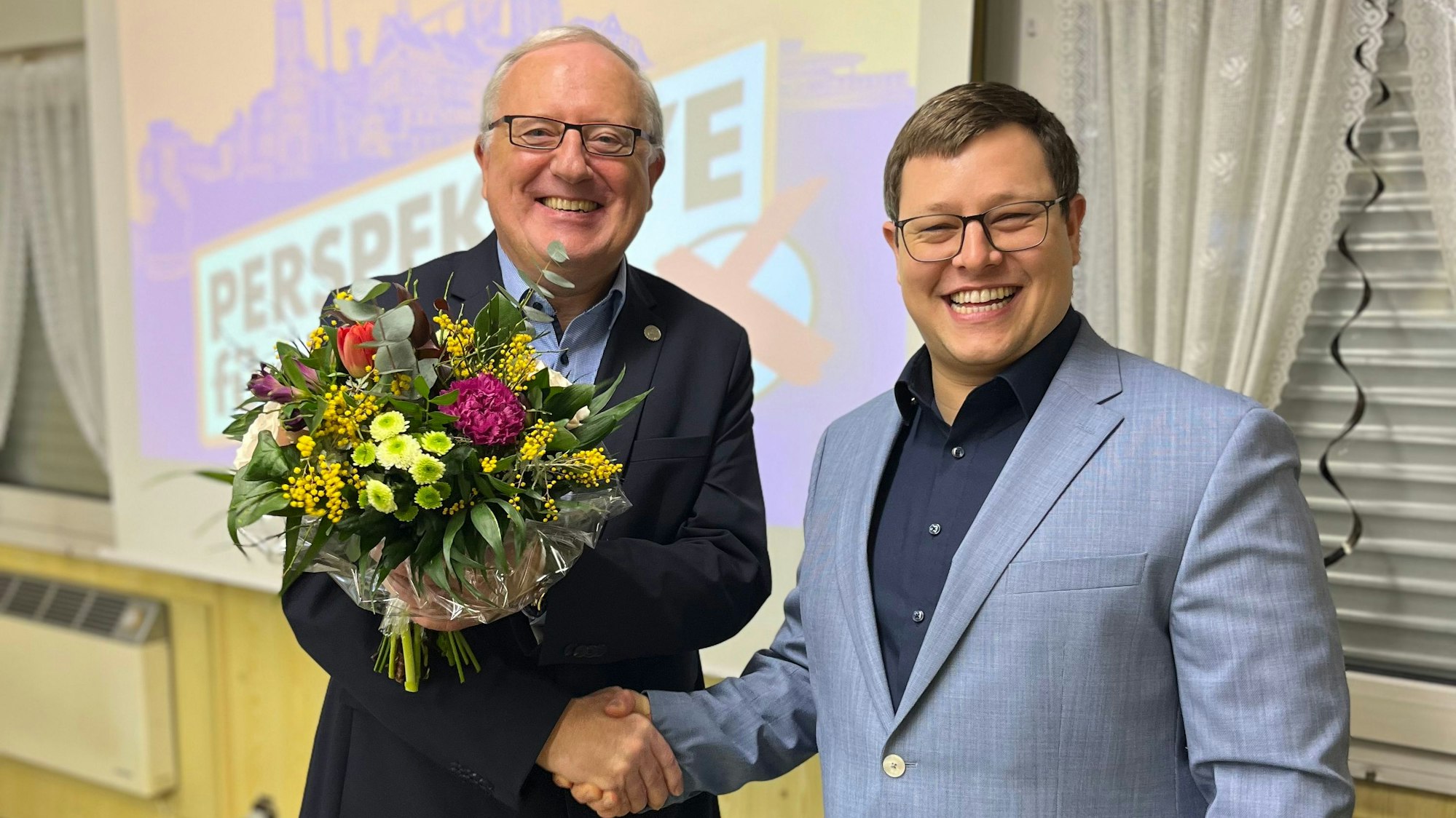 Auf dem Bild ist ein Mann zu sehen, der mit Handschlag und einem Blumenstrauße einem anderen Mann gratuliert.