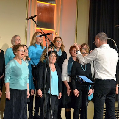Frauen stehen auf einer Bühne und singen, den Dirigenten sieht man von hinten.